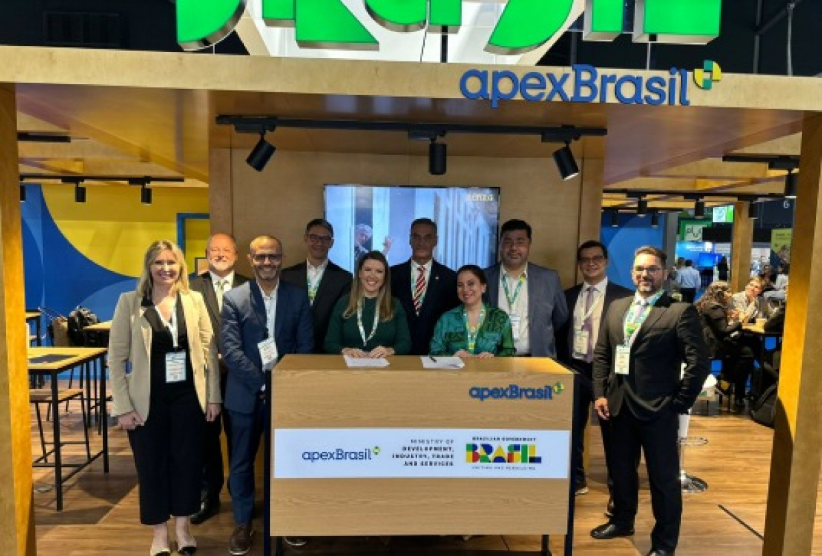 A imagem mostra representantes em um estande da ApexBrasil durante um evento internacional, promovendo o Brasil e suas oportunidades de negócios.