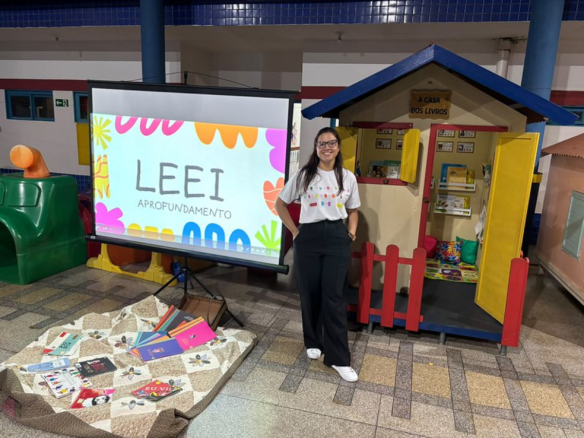 Mulher posa ao lado de uma exposição da LEEI, com livros e uma casinha temática de leitura infantil.