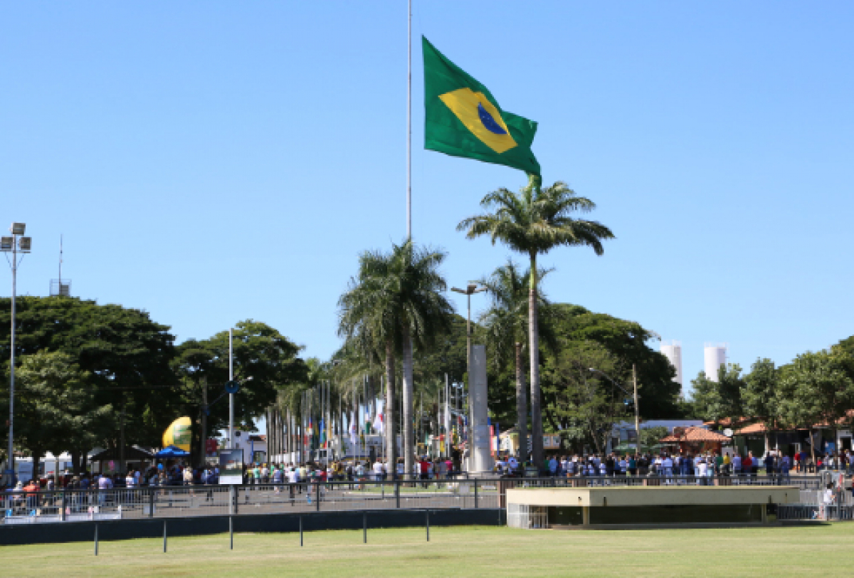 A imagem mostra uma bandeira do Brasil hasteada a meio-mastro em um espaço público com árvores, uma área gramada e uma multidão ao fundo.