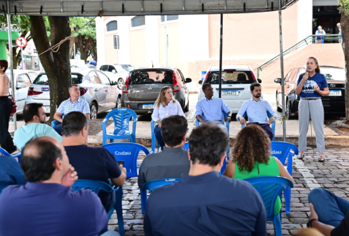 uma reunião pública ou comunitária. Todos estão sentados em cadeiras de plástico azuis marcadas com Codau.