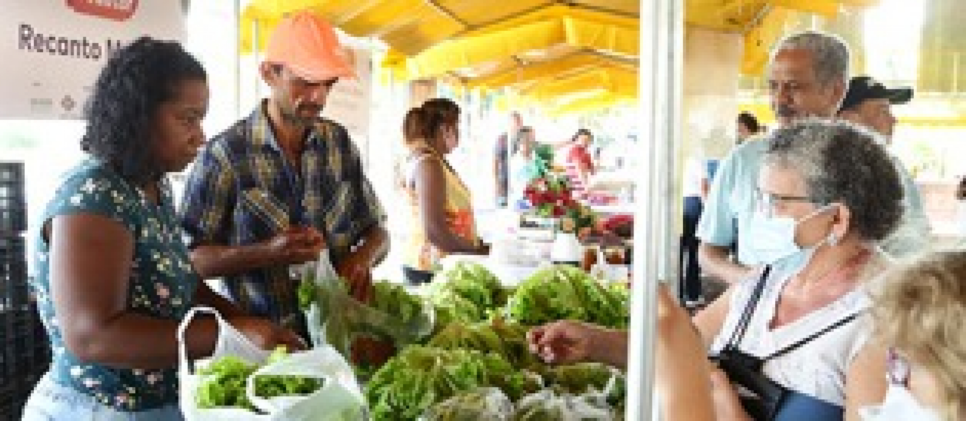 Feirantes atendem clientes em barraca de feira, oferecendo produtos agrícolas frescos, como verduras e legumes.