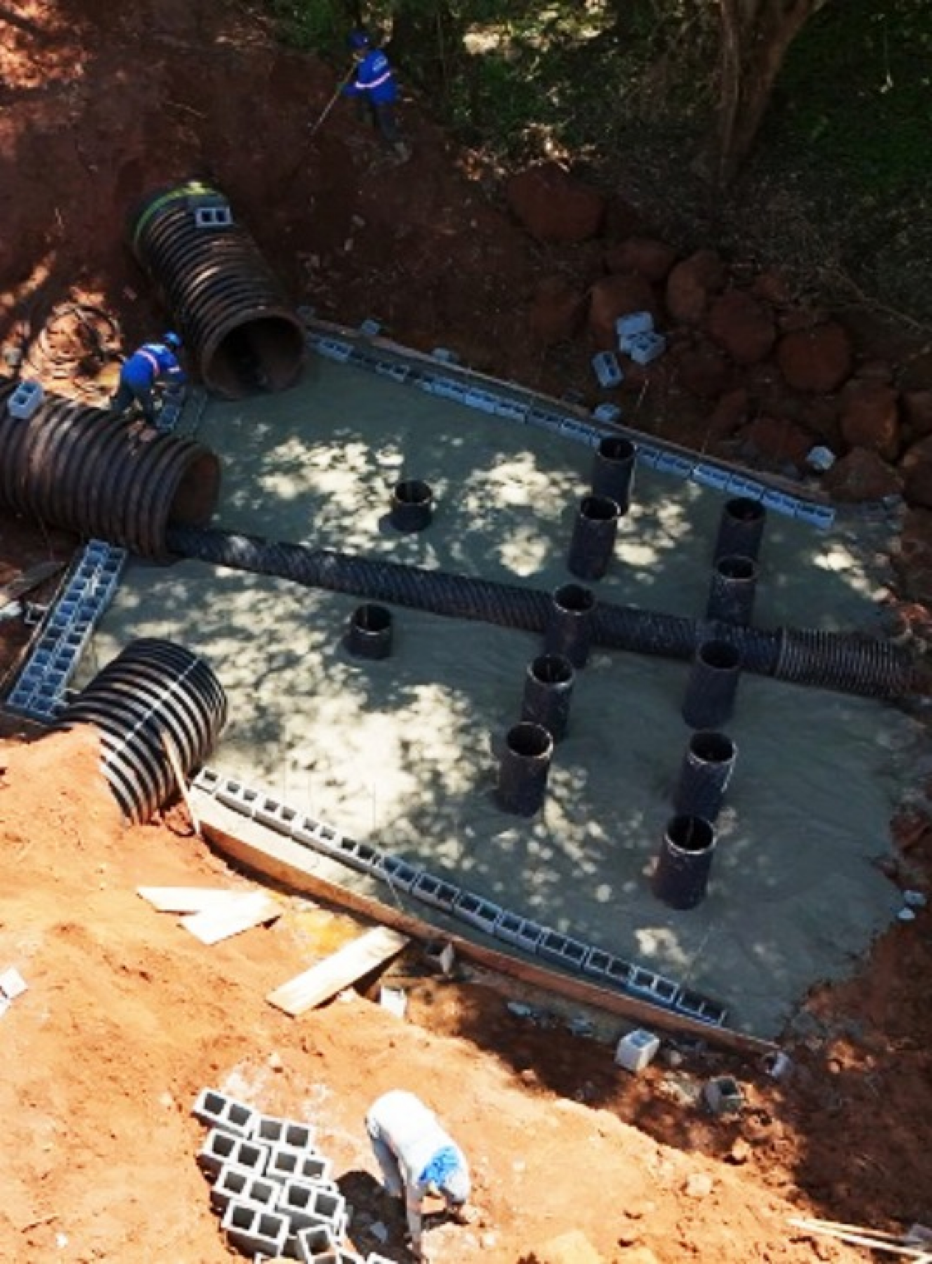 Obra de infraestrutura em andamento, com trabalhadores instalando tubulações e estruturas de concreto em uma área escavada