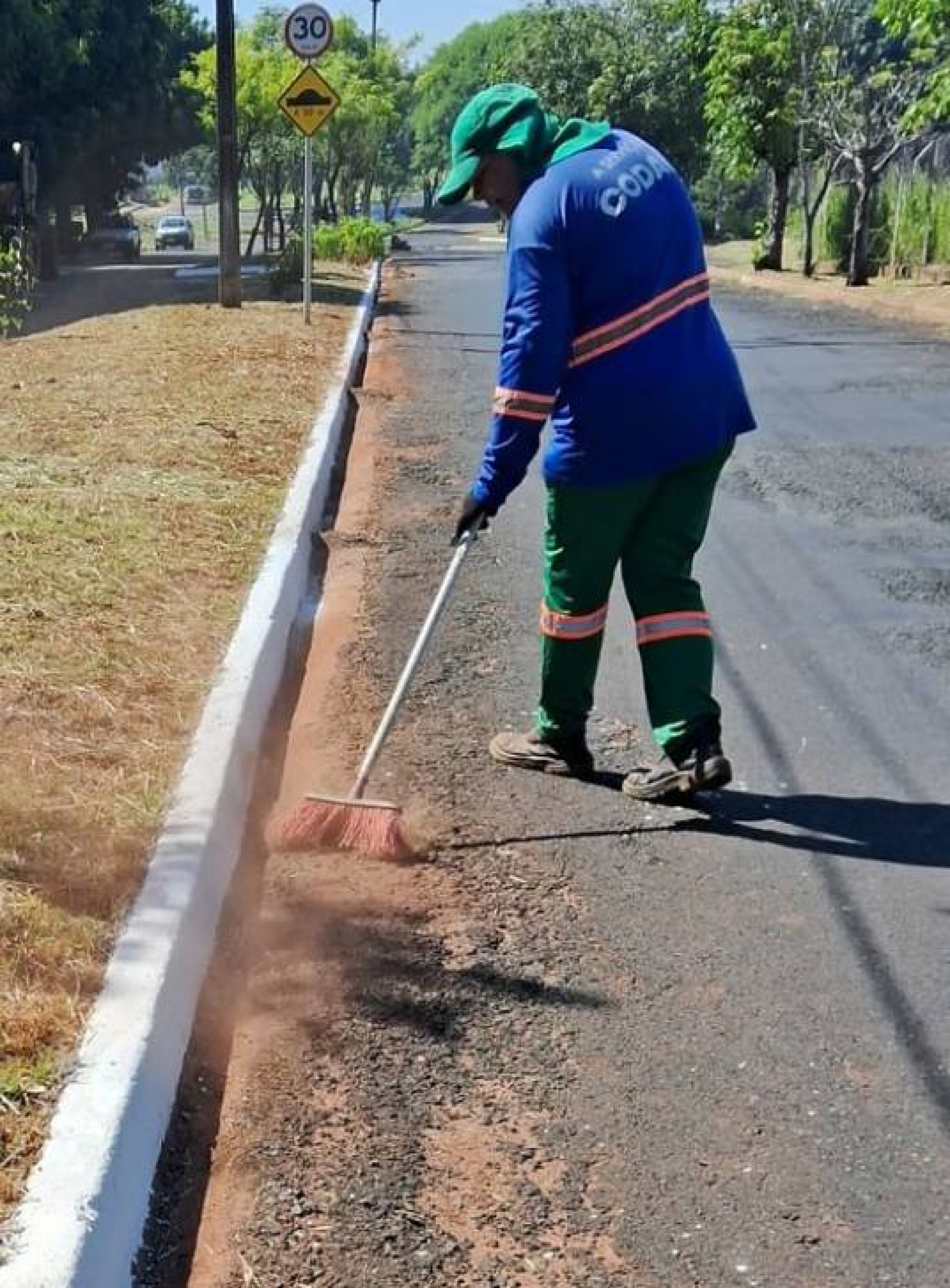 A imagem mostra uma pessoa varrendo a beira de uma rua usando um uniforme azul e verde e chapéu verde.