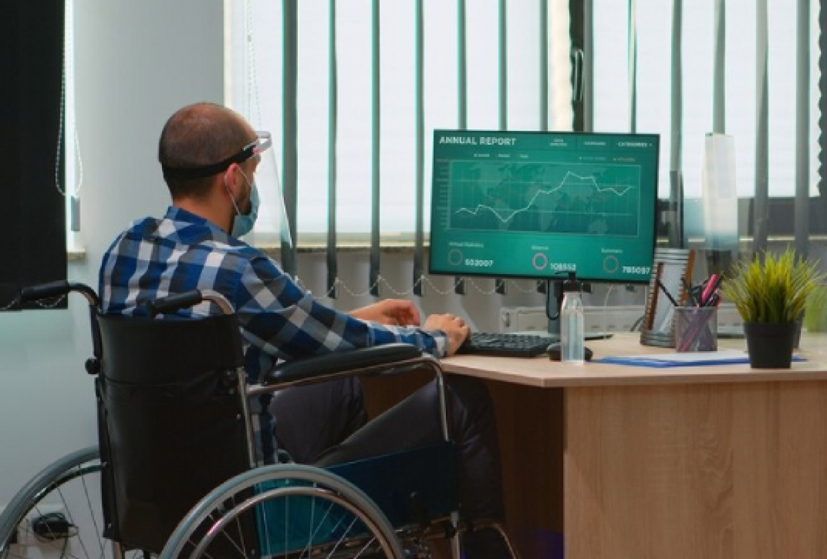 A imagem mostra uma pessoa em uma cadeira de rodas trabalhando em uma mesa, olhando para um monitor de computador. 