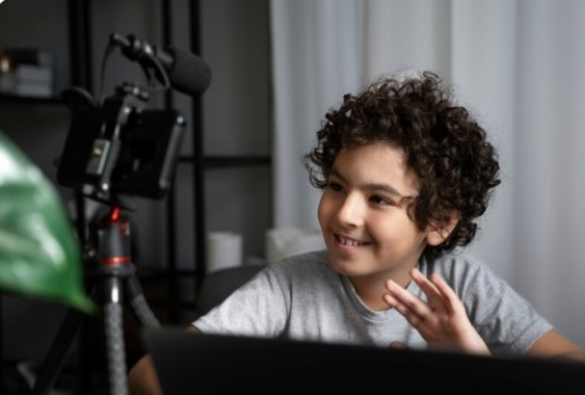 Criança sorridente grava vídeo em casa diante de câmera e microfone, em ambiente de produção audiovisual.