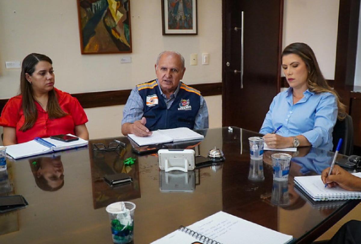 #ParaTodosVerem: Três pessoas estão reunidas ao redor de uma mesa, discutindo e fazendo anotações. Um homem de colete azul lidera a conversa, enquanto duas mulheres, uma de blusa vermelha e outra de azul, escutam atentamente. Na mesa, há itens como cadernos, canetas e copos d'água. Ao fundo, vê-se uma porta e alguns quadros.