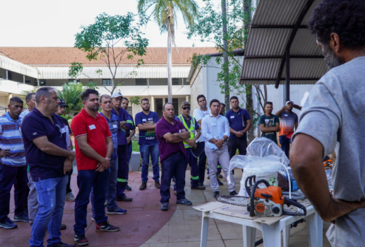 A imagem mostra um grupo de pessoas reunidas ao ar livre, assistindo a uma demonstração ou treinamento, com equipamentos como uma motosserra em uma mesa no centro.