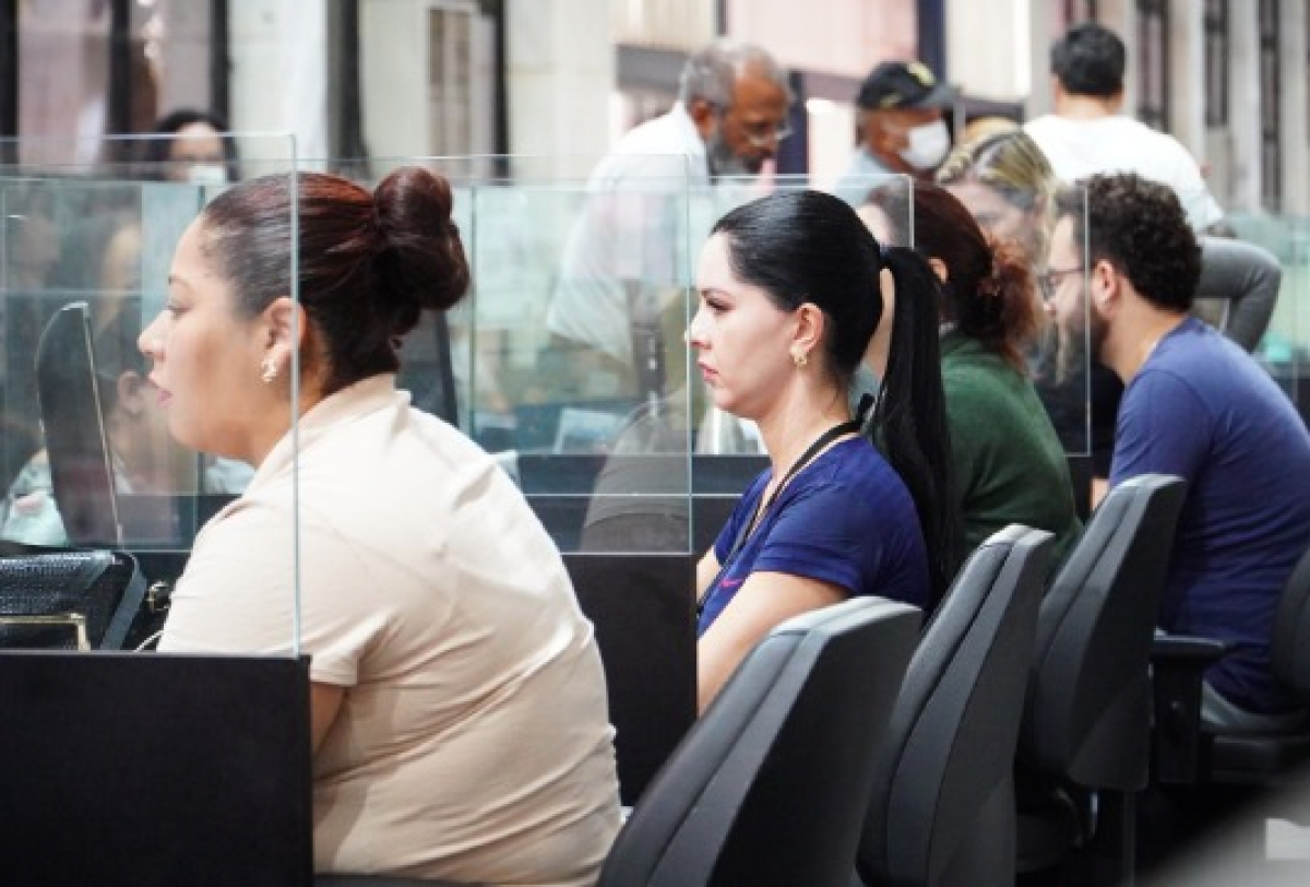 Atendentes prestam serviços ao público em balcões de atendimento com divisórias de acrílico. 