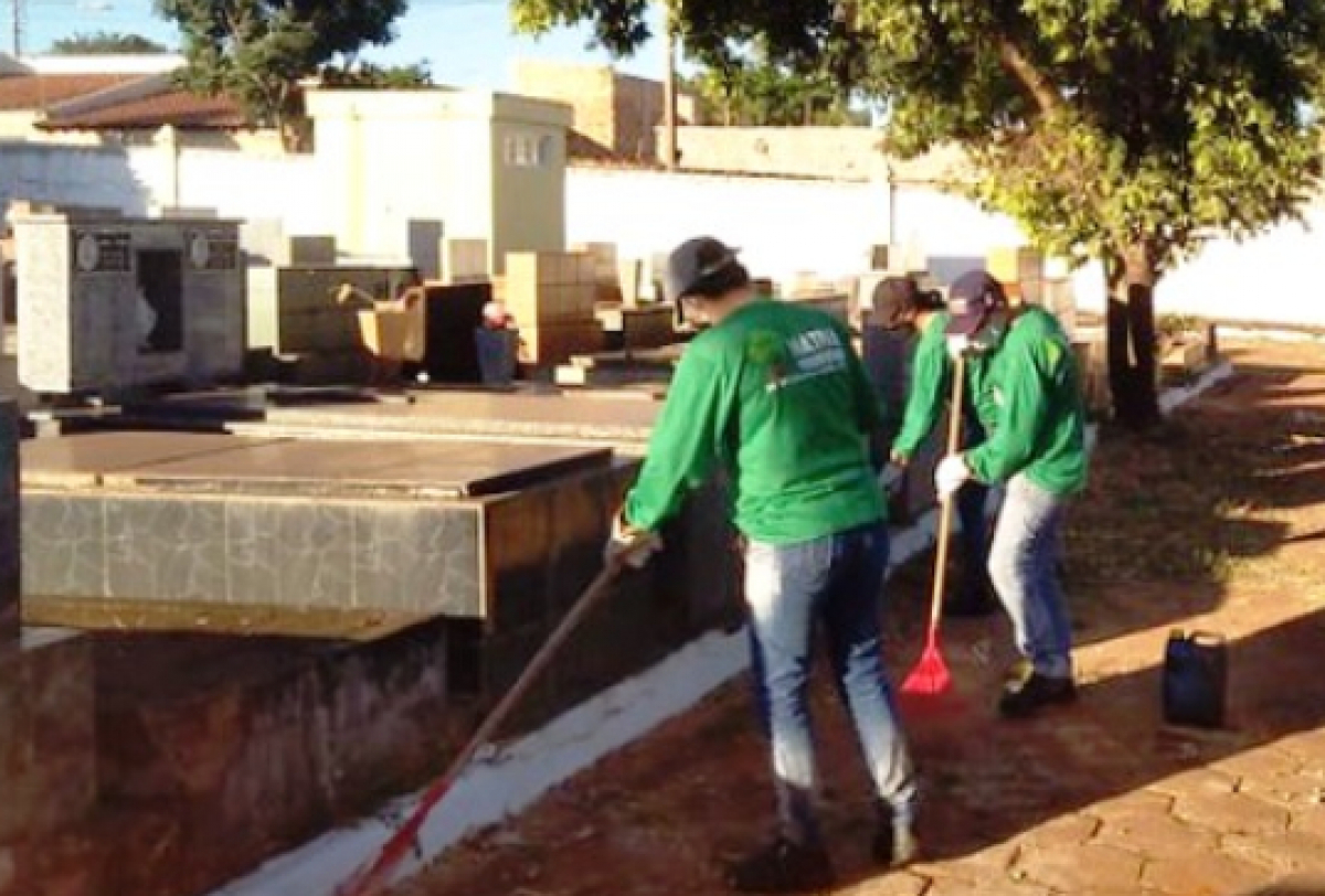 Funcionários realizam serviço de limpeza e manutenção em cemitério, varrendo e cuidando do espaço entre os túmulos.