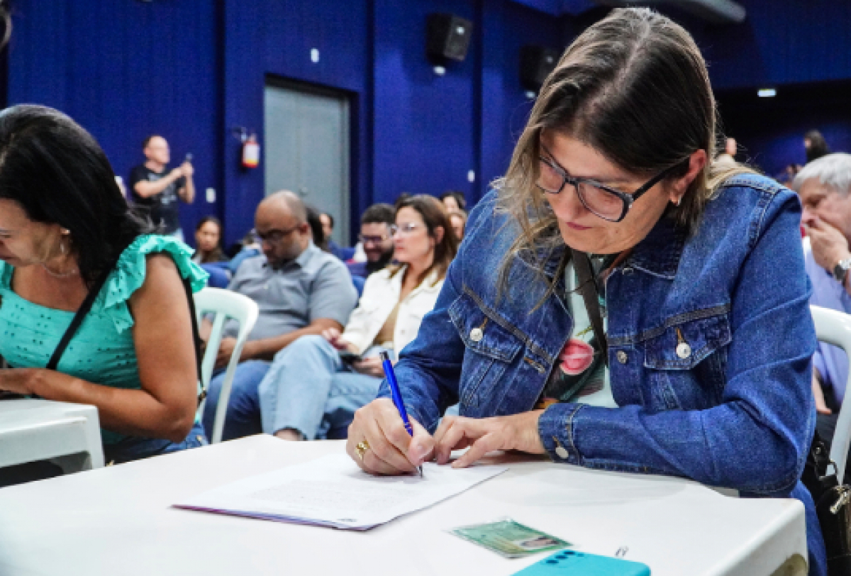 Mulher preenche formulário durante evento em auditório, com outras pessoas atentas ao fundo.
