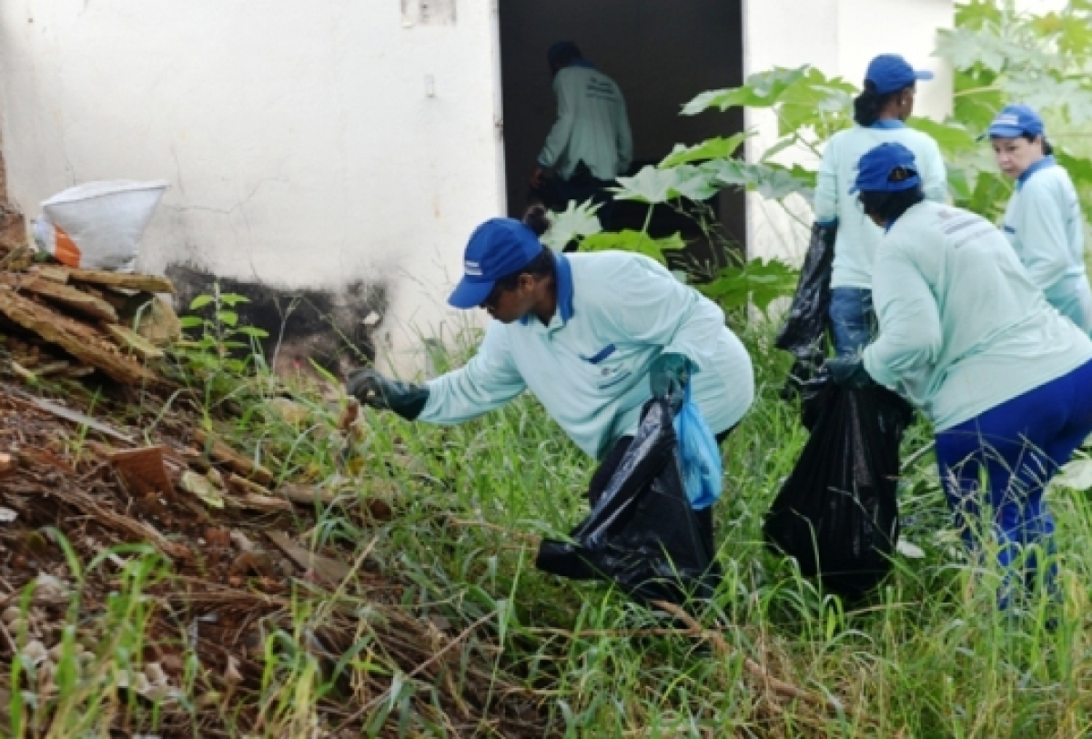 A imagem mostra um grupo de pessoas participando de uma atividade de limpeza, vestindo camisetas azuis claras e bonés azuis, coletando lixo em sacos pretos. O local tem vegetação e há um prédio branco ao fundo. 