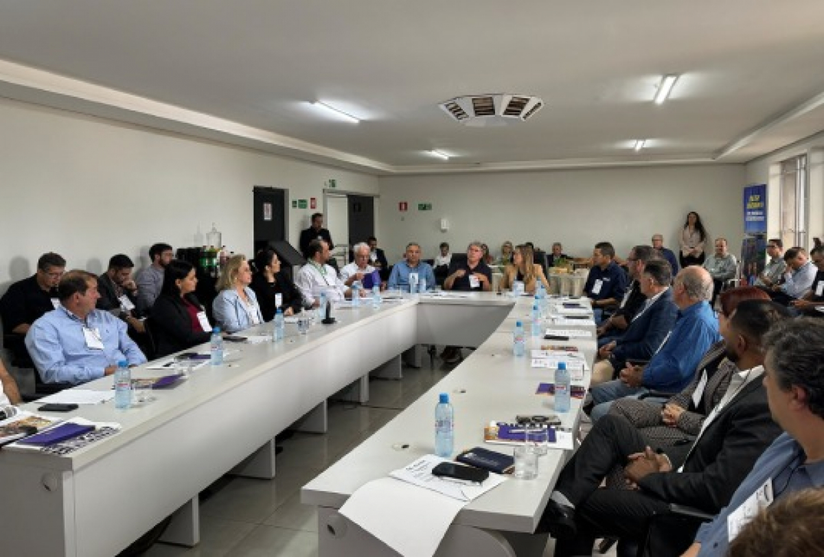 Reunião formal com diversas pessoas em torno de mesas em U, discutindo assuntos institucionais.