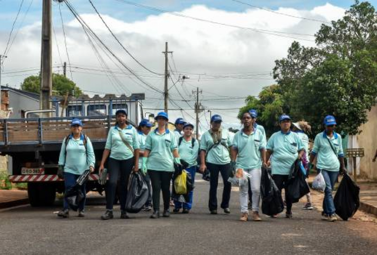 A imagem mostra um grupo de pessoas caminhando por uma rua, vestindo camisetas azuis claras e bonés azuis, carregando sacos de lixo. Um caminhão está estacionado ao lado. 