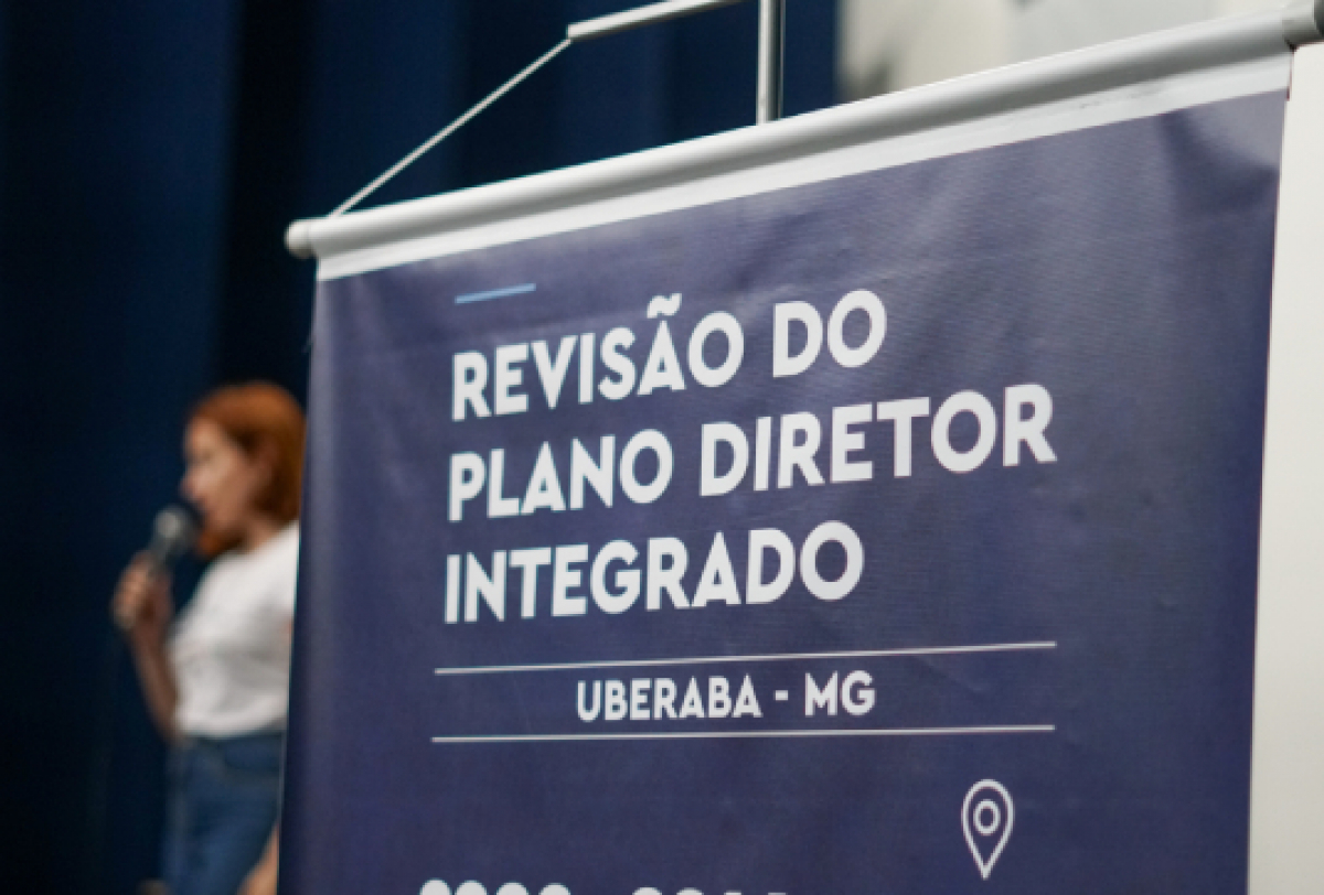 Banner anuncia a revisão do Plano Diretor Integrado de Uberaba-MG, com uma palestrante ao fundo.