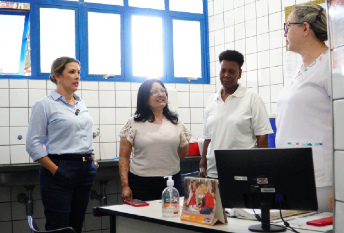 Profissionais conversam em ambiente de unidade de saúde ou escola, possivelmente discutindo ações ou melhorias no atendimento.