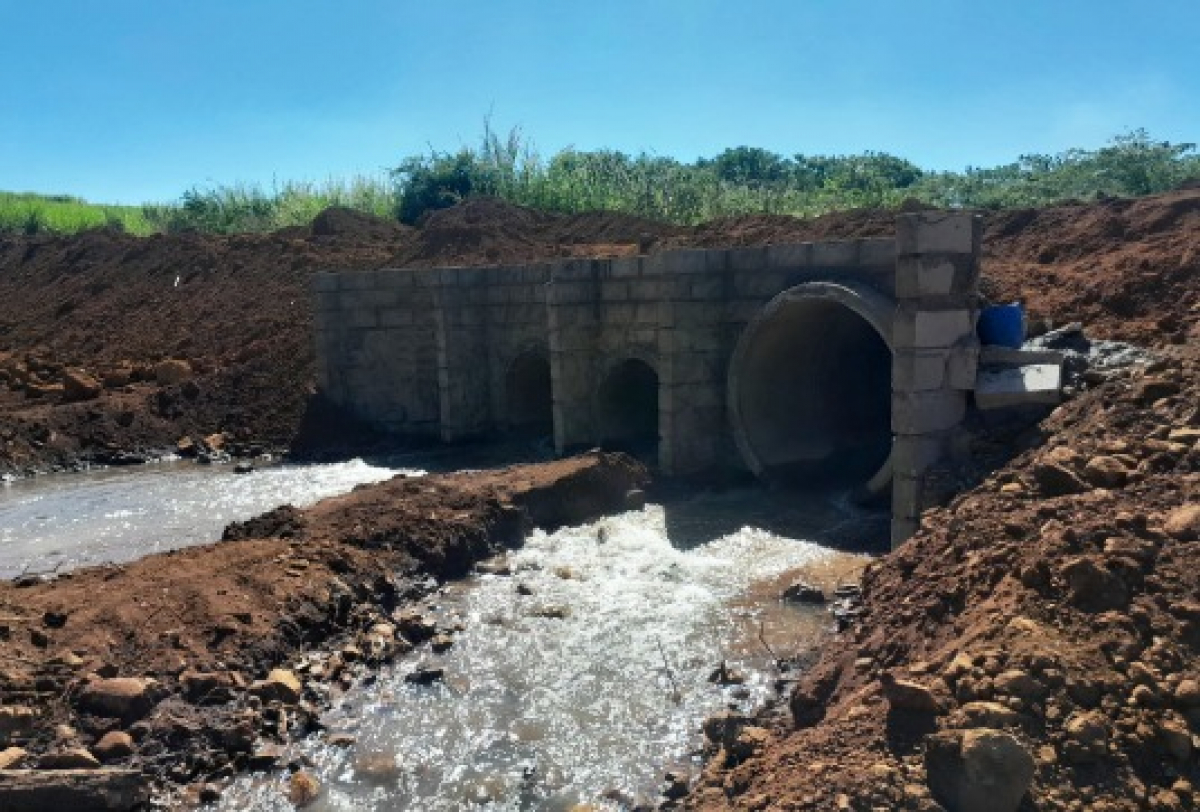 Estrutura de drenagem com tubos de concreto e paredes de alvenaria canaliza o fluxo de água em área rural.