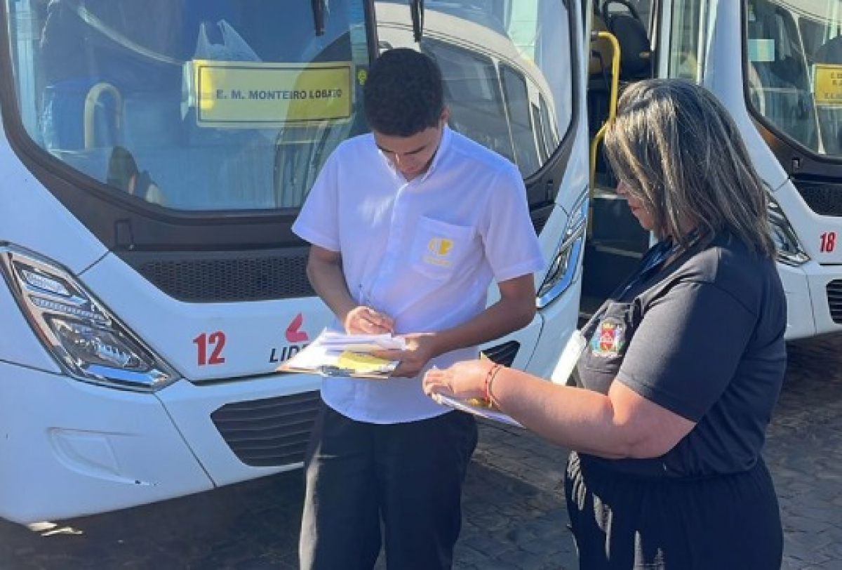 Funcionários realizam conferência de documentos em frente a ônibus escolares estacionados.