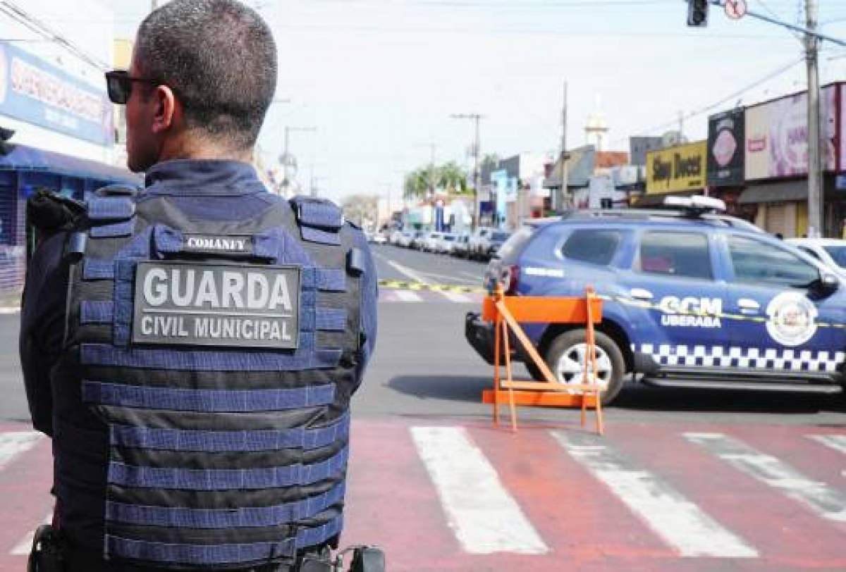 A imagem mostra um agente da Guarda Civil Municipal em frente a uma área isolada, com uma viatura ao fundo.