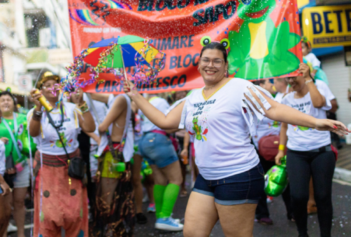 É uma cena vibrante de carnaval, com participantes usando camisetas brancas com um gráfico de sapo e texto BLOCO SAPUCAIA.