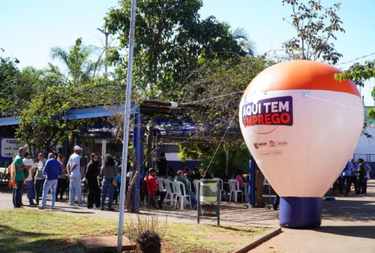 Evento de empregabilidade reúne pessoas em fila e cadeiras sob estrutura com balão informativo Aqui Tem Emprego.