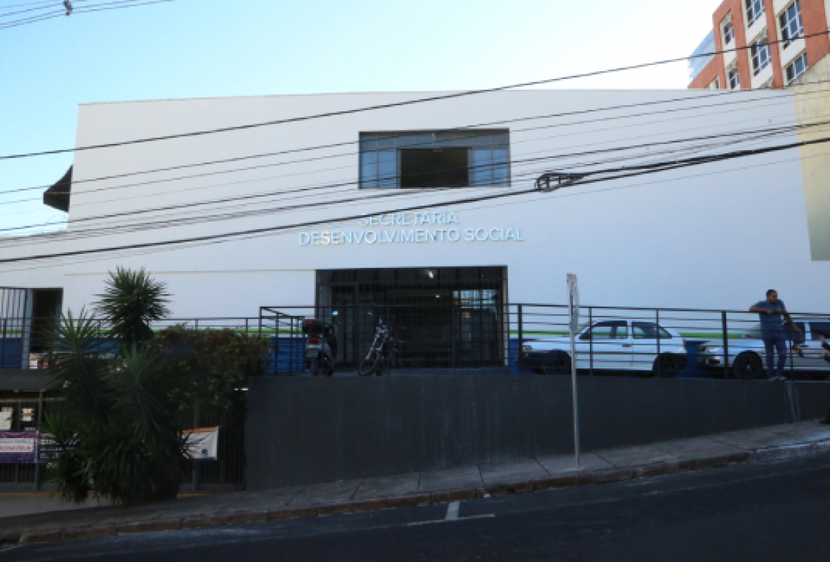 A imagem mostra a fachada de um edifício com a inscrição SECRETARIA DESENVOLVIMENTO SOCIAL na entrada, com veículos estacionados em frente.