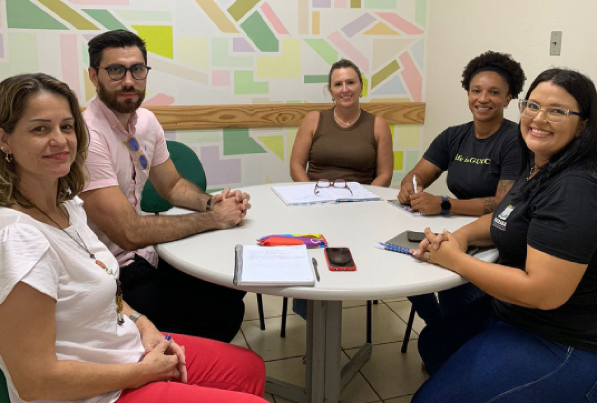 Cinco pessoas sentadas ao redor de uma mesa em uma reunião, sorrindo para a câmera.