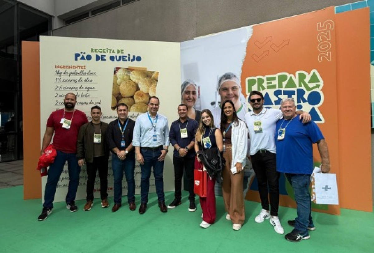 Grupo de participantes posa para foto em evento gastronômico Prepara Gastro 2025, com painel decorativo e receita de pão de queijo ao fundo.