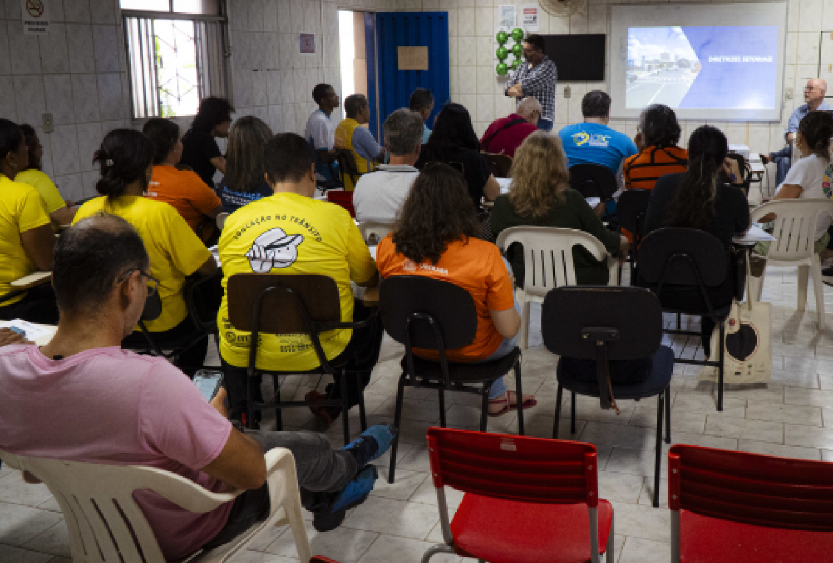 Apresentação em sala fechada com pessoas sentadas, muitas usando camisetas de organizações ou eventos.