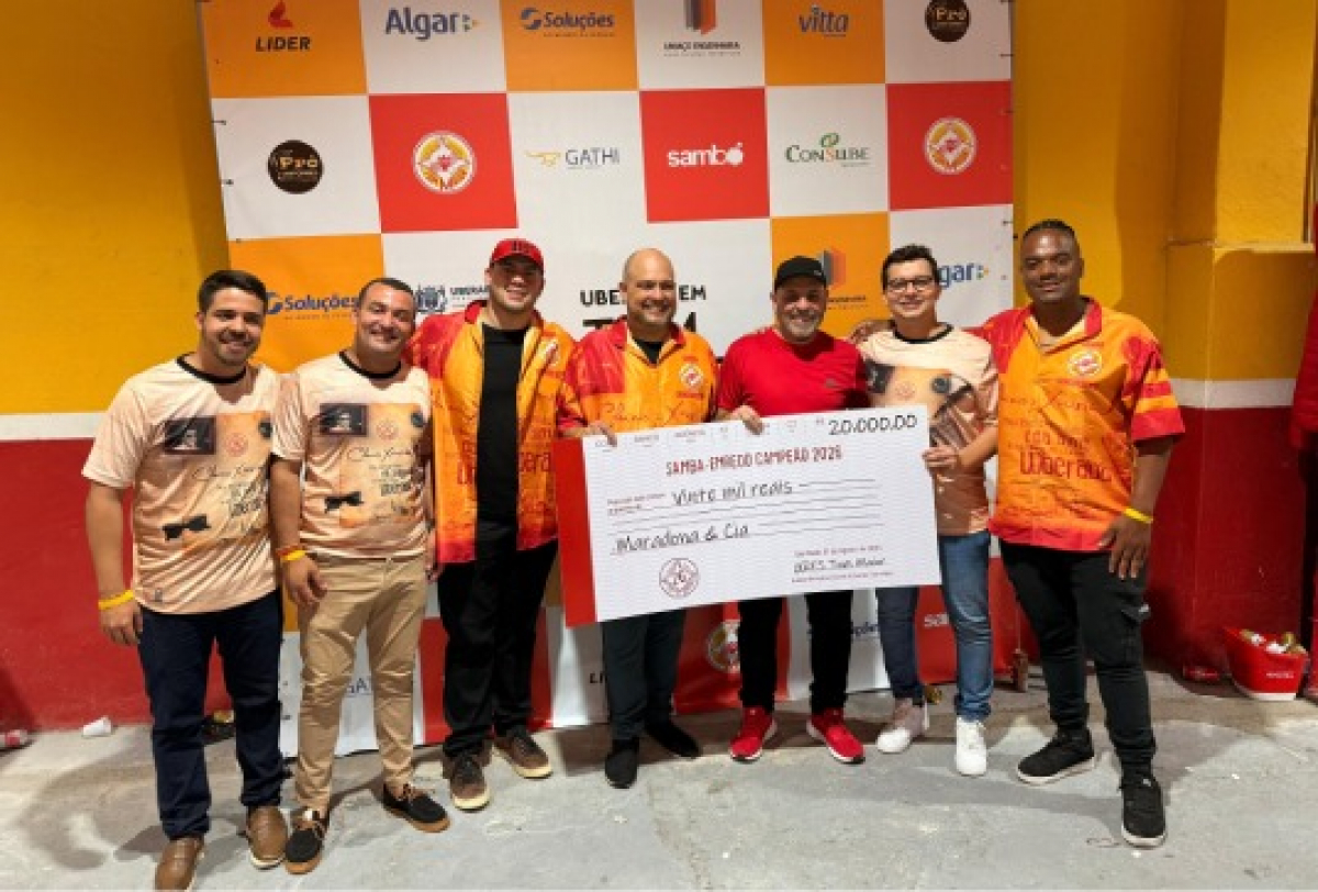 Grupo de pessoas posa sorridente segurando cheque simbólico de premiação em evento esportivo ou cultural.