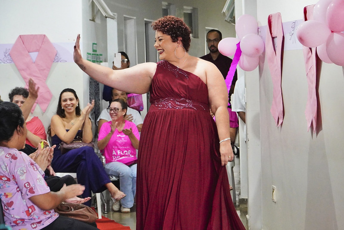 Mulher em vestido vermelho é recebida com aplausos em evento decorado com laços e balões cor-de-rosa.