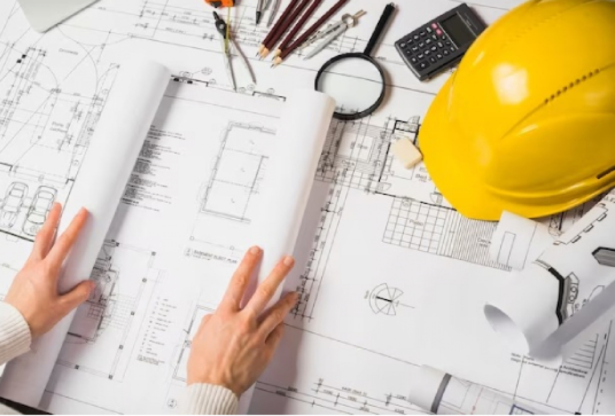 Imagem de um ambiente de trabalho de engenharia ou arquitetura, com plantas técnicas, capacete de segurança, instrumentos de desenho e uma calculadora.