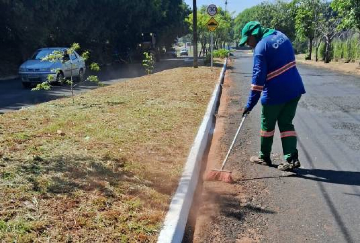 A imagem mostra uma pessoa varrendo a beira de uma rua usando um uniforme azul e verde e chapéu verde.