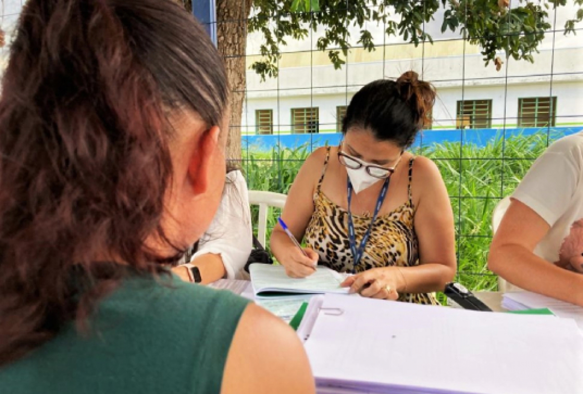 Uma pessoa escreve em um caderno enquanto um grupo de pessoas está reunido ao ar livre, possivelmente participando de uma atividade envolvente.