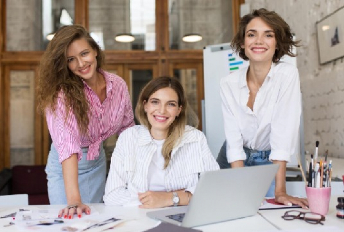 Três mulheres trabalham juntas em um escritório, reunidas ao redor de uma mesa com um notebook.