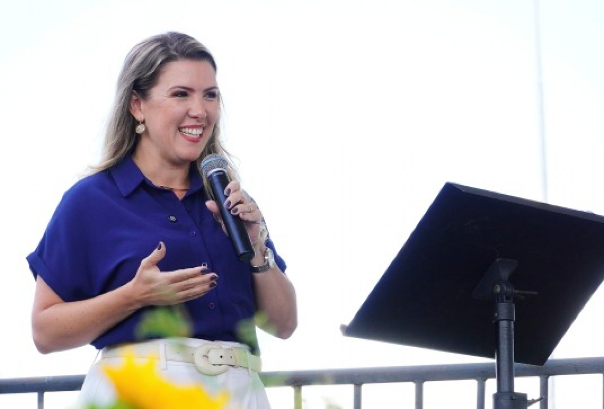 Mulher sorridente discursa ao ar livre, segurando um microfone ao lado de um pedestal.