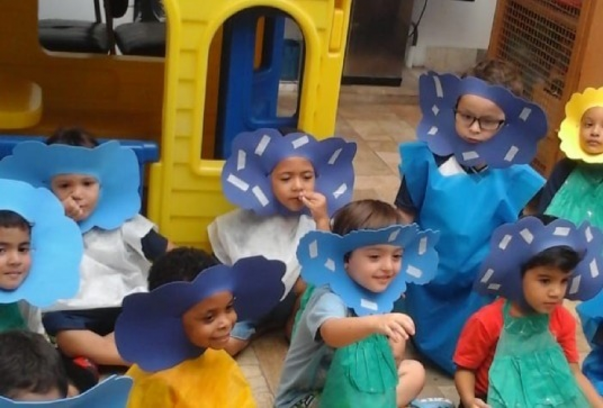 Crianças pequenas participam de uma atividade escolar fantasiadas com flores coloridas feitas de papel.