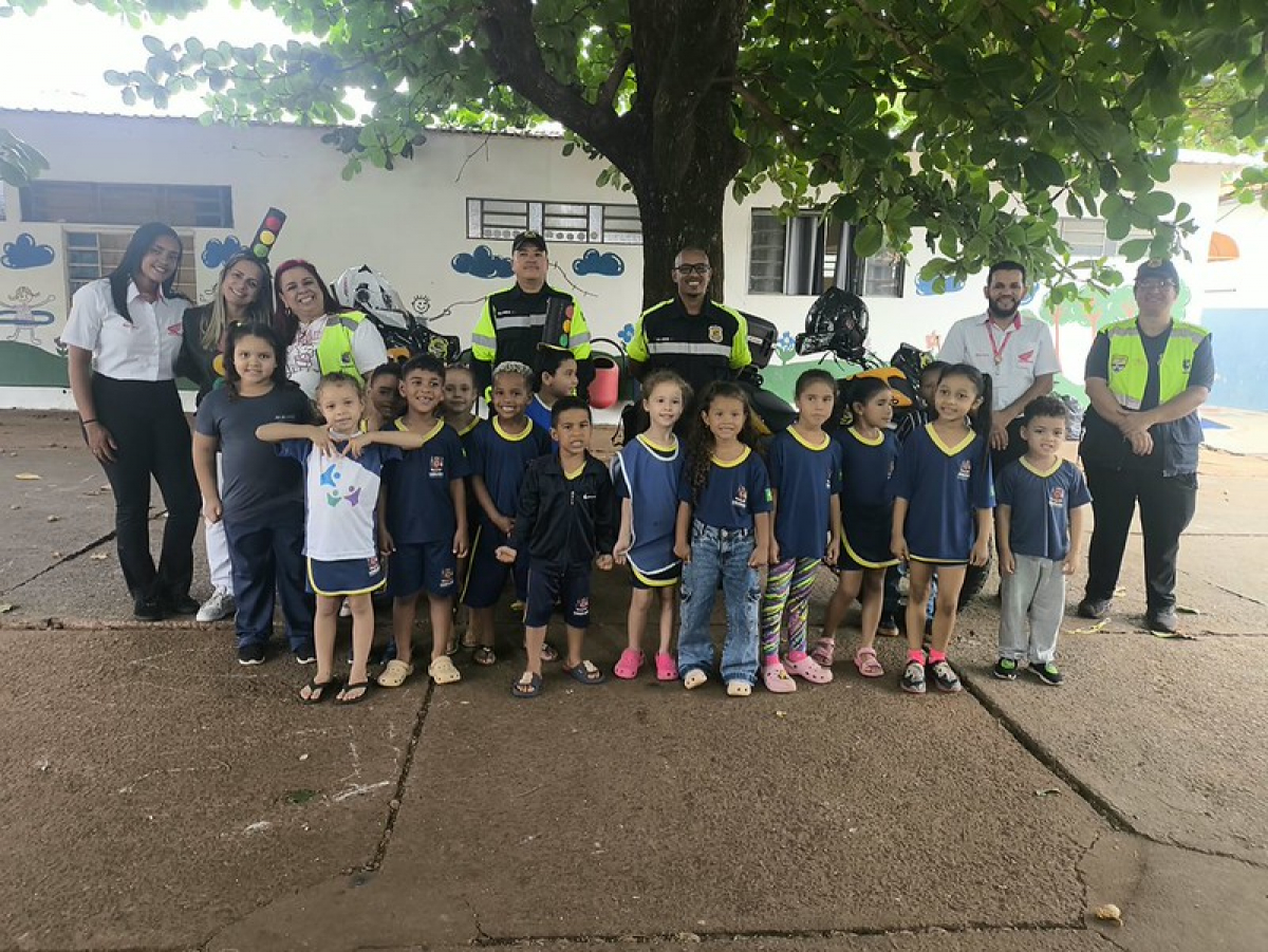 Crianças e profissionais da segurança posam juntos em frente a uma escola, durante uma atividade educativa.