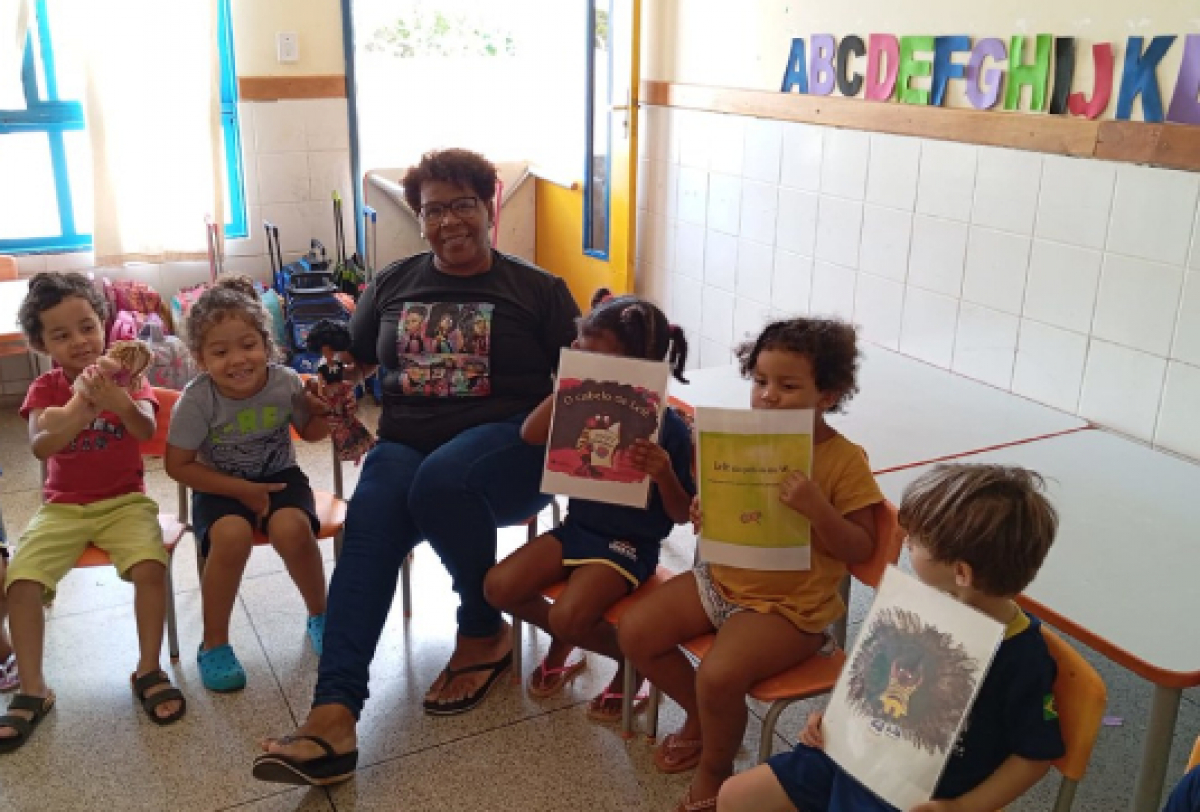 Crianças em uma sala de aula participam de uma atividade de leitura com uma professora, segurando livros e bonecas.