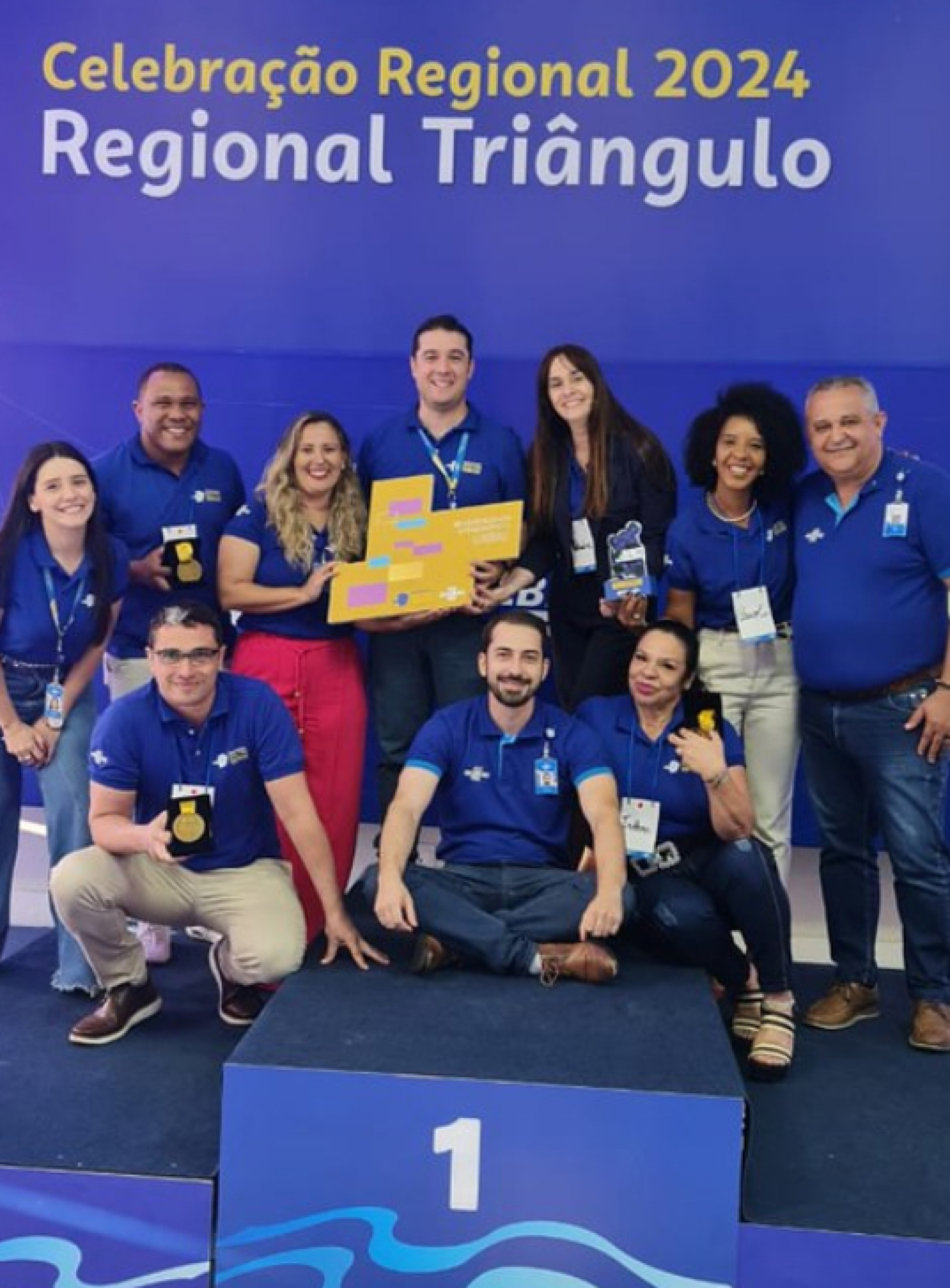 A imagem mostra um grupo de pessoas posando juntas em um pódio com um painel de fundo que lê Celebração Regional 2024 Regional Triângulo. 