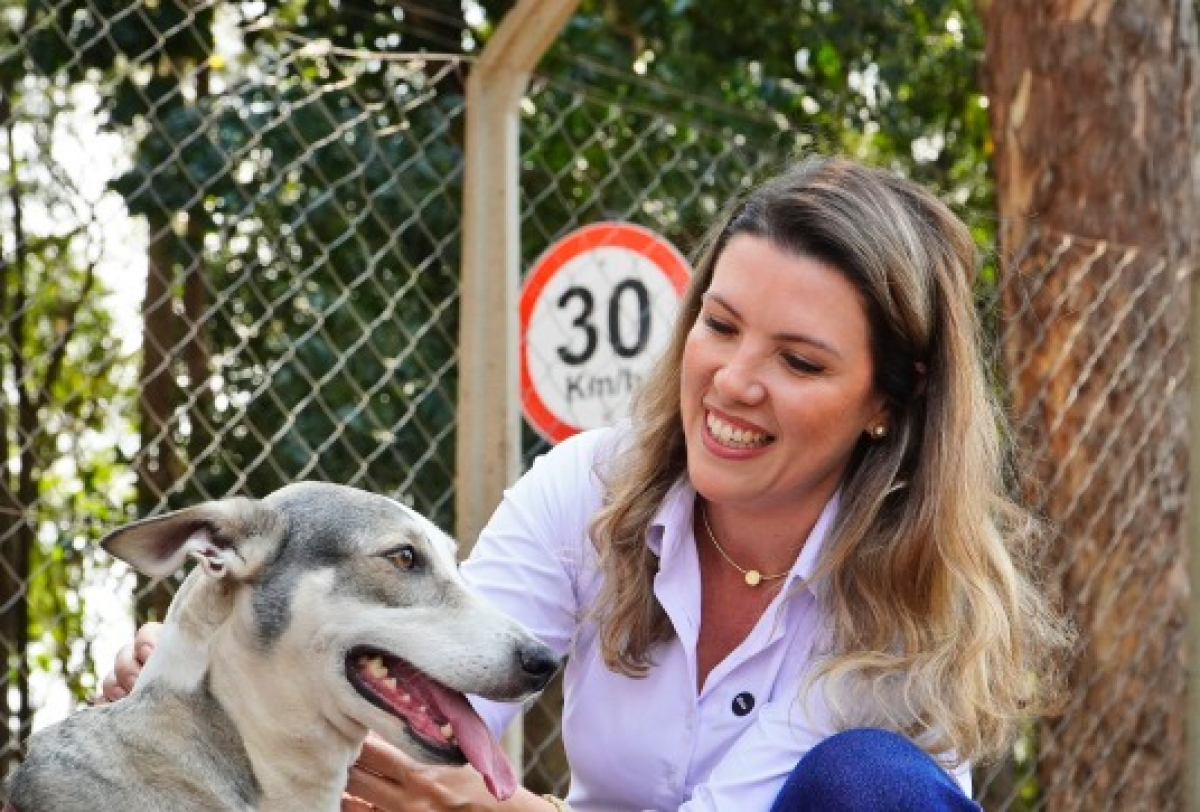 Uma mulher sorri ao lado de um cachorro em frente a uma cerca, com uma placa de limite de velocidade ao fundo.
