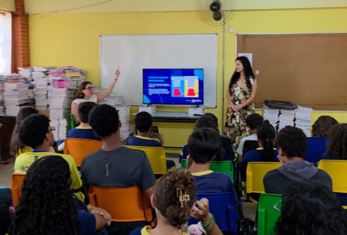 A imagem mostra uma palestra em sala de aula, com uma apresentadora explicando um slide para alunos atentos.