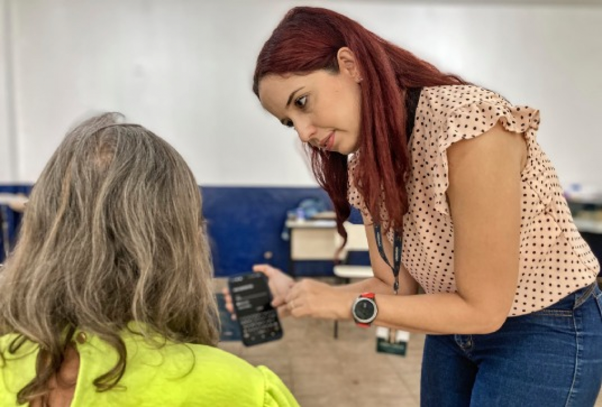 Mulher orienta uma pessoa idosa no uso do celular em ambiente de sala de aula.