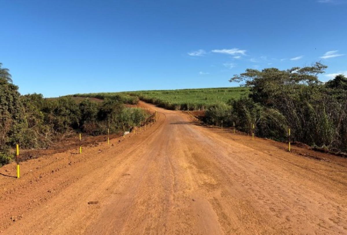 Estrada de terra em área rural cercada por vegetação, sob céu limpo e ensolarado.