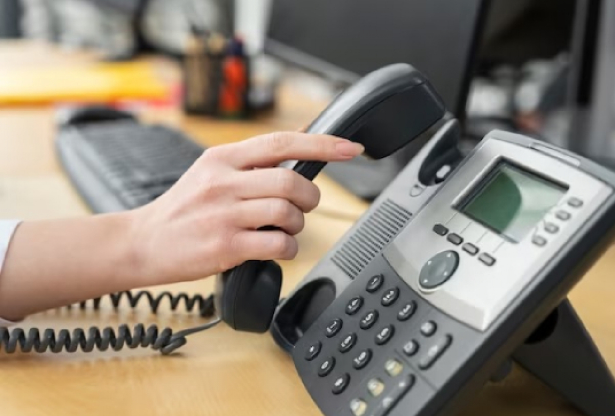 A imagem mostra a mão de uma pessoa segurando o receptor de um telefone de mesa em um ambiente de escritório.