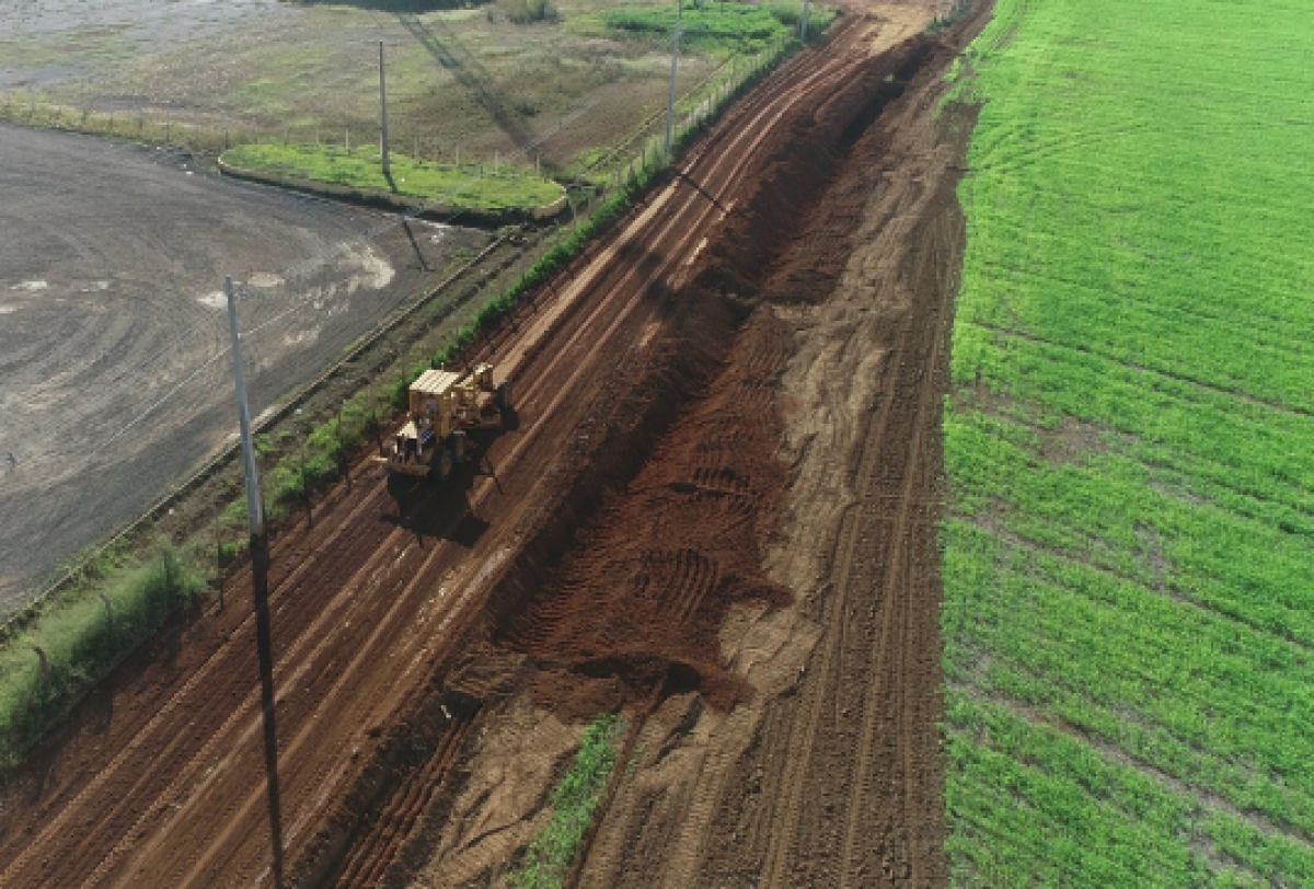 Imagem mostra obra de terraplanagem com máquina niveladora entre área agrícola e terreno compactado.