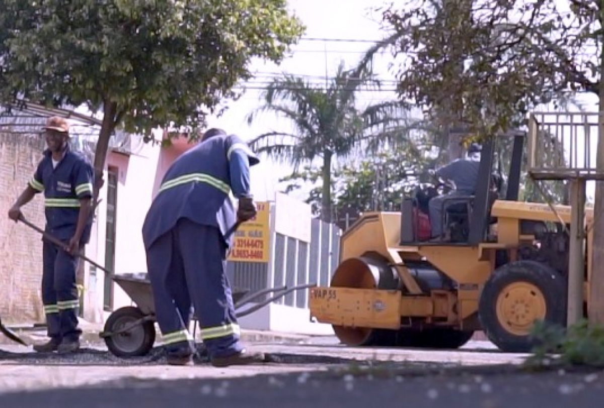 Operação de tapa-buracos com equipe e rolo compactador realizando manutenção asfáltica em via urbana.