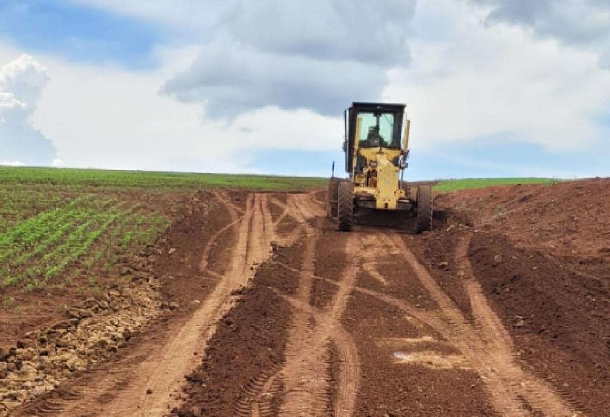 A imagem mostra uma máquina niveladora amarela trabalhando em uma estrada de terra em uma área rural. 