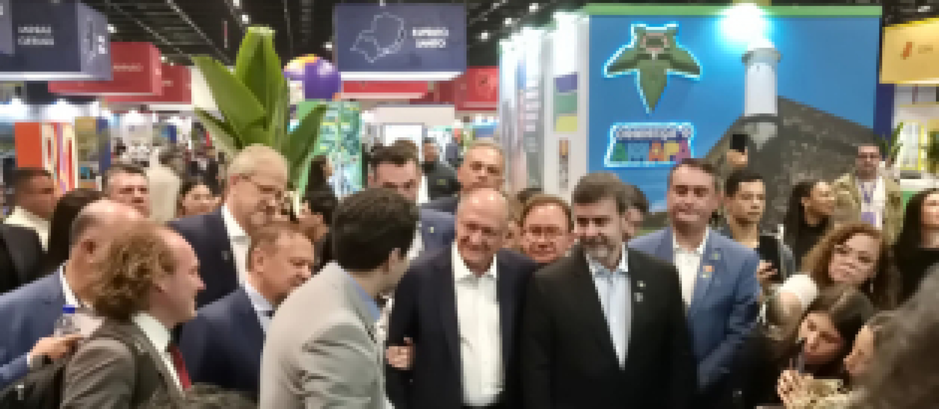 Grupo de autoridades e visitantes reunidos em um estande de feira ou evento com foco em estados brasileiros.
