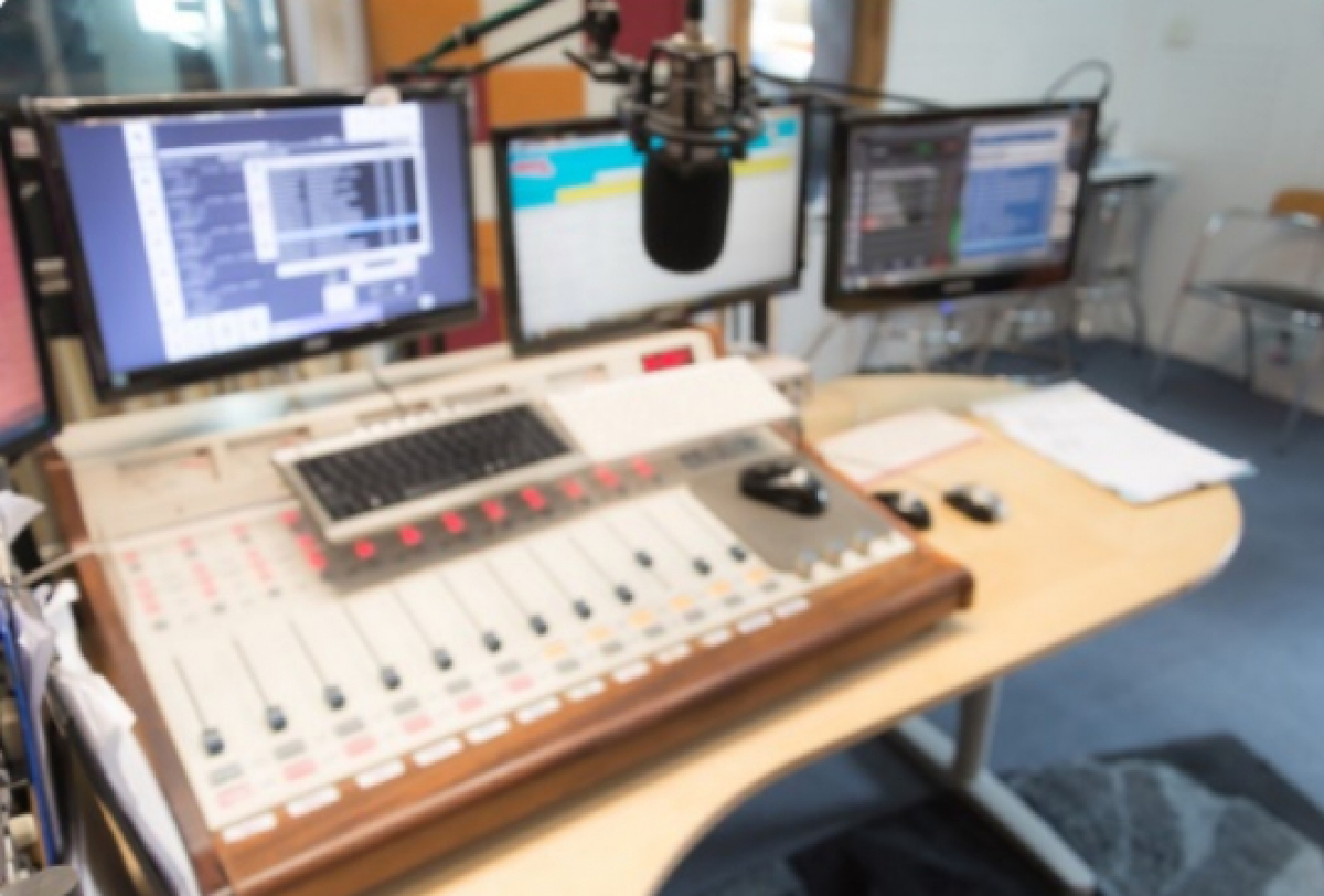 Estúdio de rádio com mesa de som, microfone e múltiplas telas ligadas em operação.