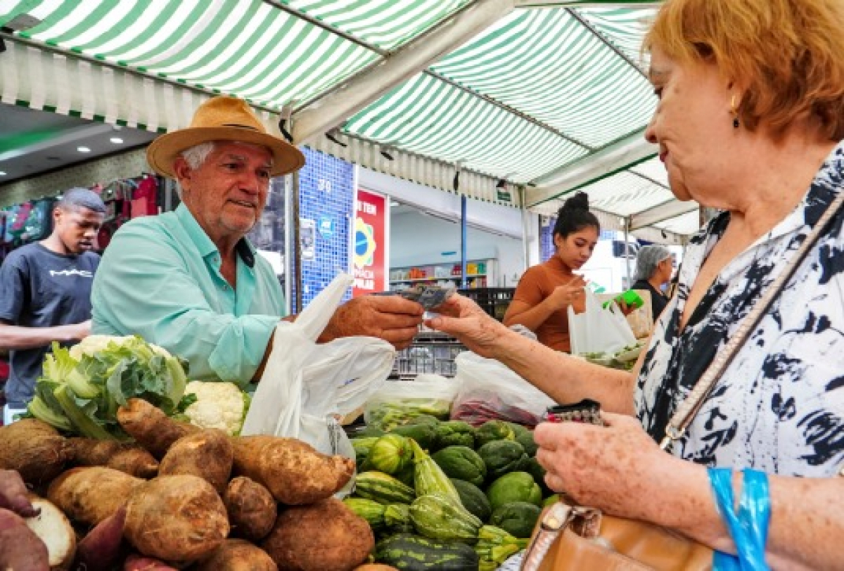 Feirante entrega produto a cliente em uma barraca de frutas e verduras.